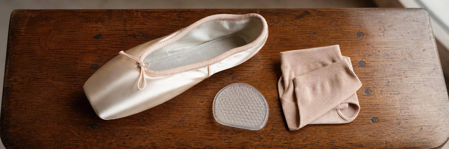 Overhead flat lay on a warm wood bench: a single satin pointe shoe on its side next to a gel toe pad and a folded pair of pointe socks, the shoe positioned as if being examined for fit.