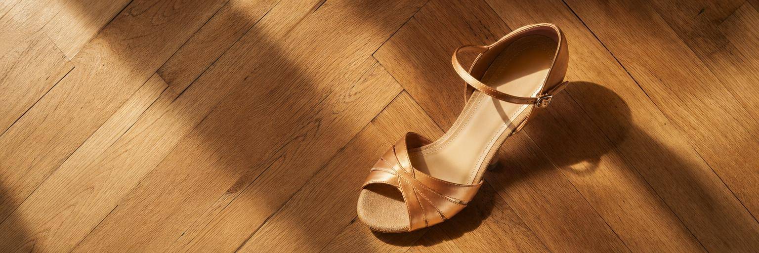 Overhead flat lay on a warm parquet floor: a single tan open-toe Latin dance heel with a slim ankle strap, photographed to show the open toe and suede sole, the matching shoe just out of frame.