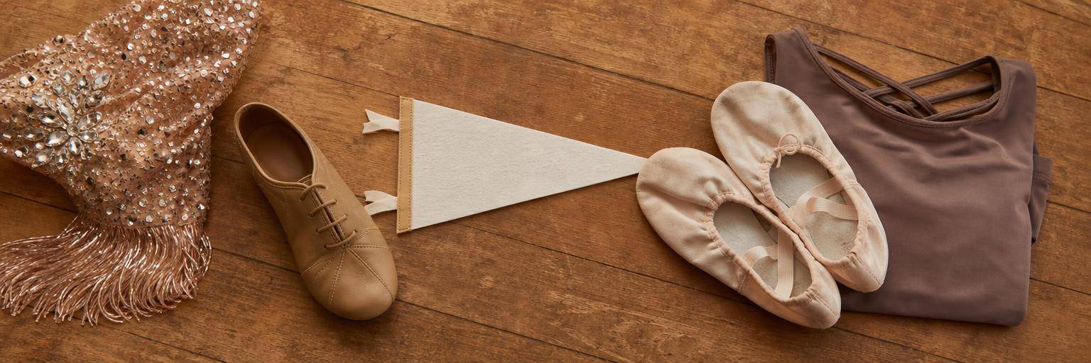 An overhead flat lay on a warm wood studio floor contrasting two dance worlds side by side: on one side a sparkly competition-style accessory and a tan jazz shoe, on the other a soft ballet slipper and a folded modern-dance practice top, with a college pennant resting between them to suggest the bridge from competition to college dance.