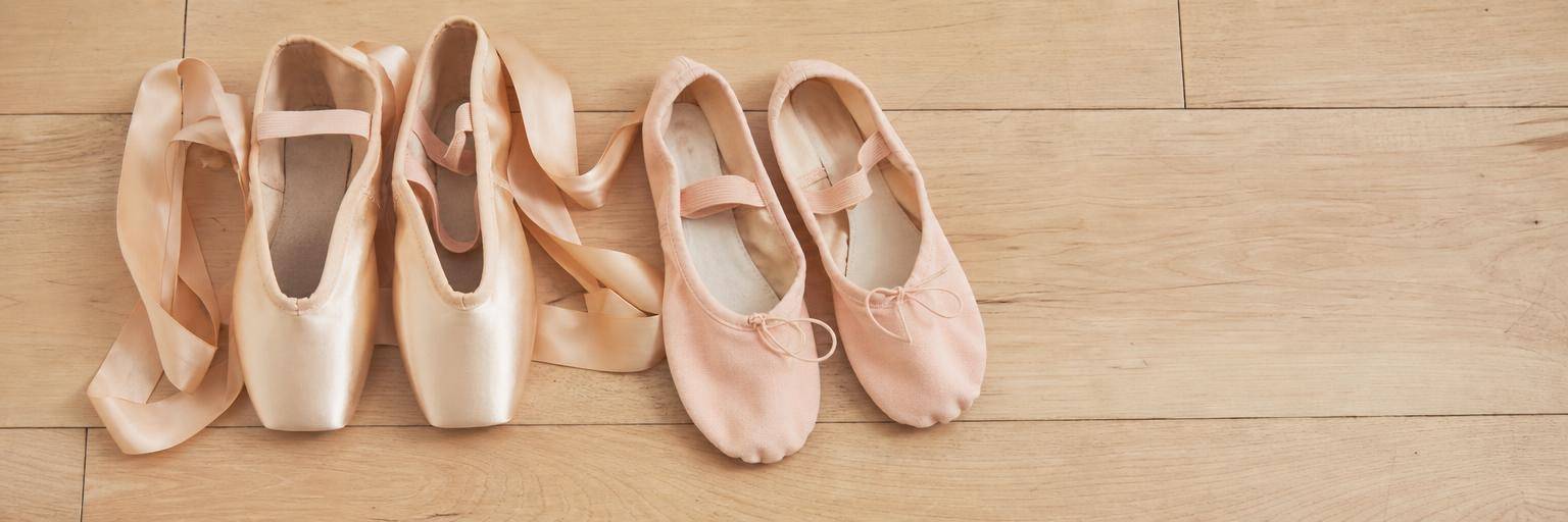 Overhead flat lay on a pale wood ballet studio floor: a pair of soft demi-pointe shoes with a pointe-shoe shape but a soft sole and loosely coiled ribbons, set beside a pair of regular pink ballet slippers to show they are different shoes.