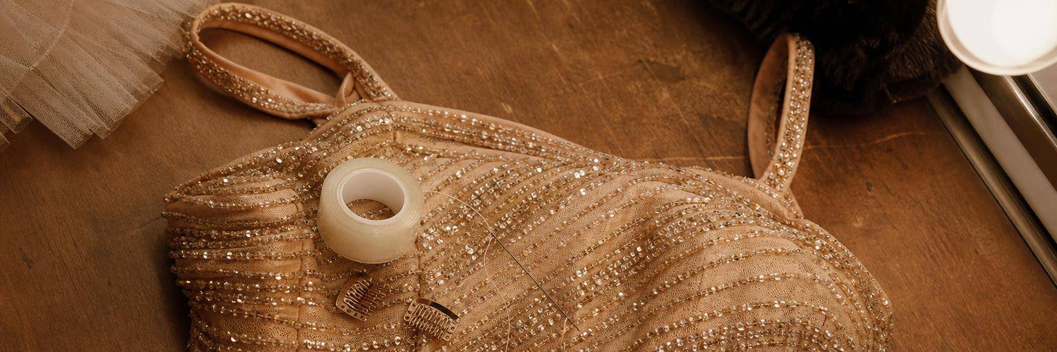 Overhead flat lay on a warm wood dressing-room counter: a sequined costume bodice laid flat with a small roll of clear body tape, a few toupee clips, and a threaded needle resting on it, the backstage fixes for a slipping strap.