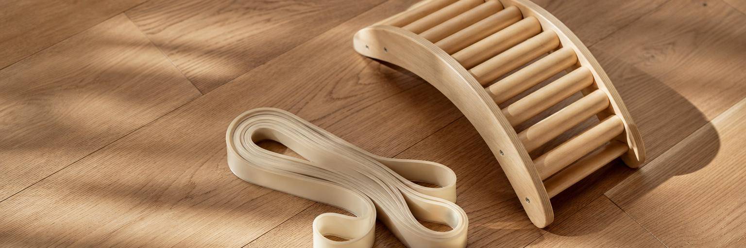Overhead flat lay on a warm wood floor: a coiled elastic resistance band in the foreground beside a wooden arch-stretcher device set slightly back, the band framed as the safer, simpler choice.